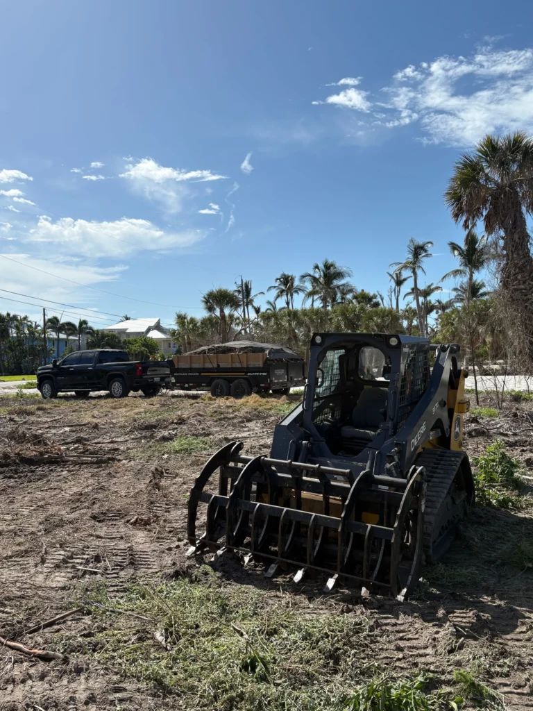 John Deere skid steer with a root grapple clearing a coastal lot with a loaded debris trailer staged for haul-off in Southwest Florida