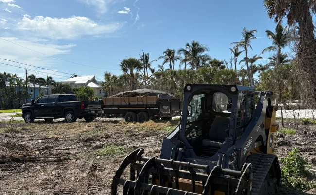 John Deere skid steer with a root grapple clearing a coastal lot with a loaded debris trailer staged for haul-off in Southwest Florida